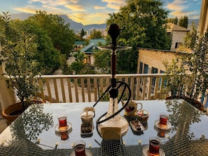 A glass table with a hookah, tea glasses, and a teapot on a balcony overlooking a residential area with trees and buildings.