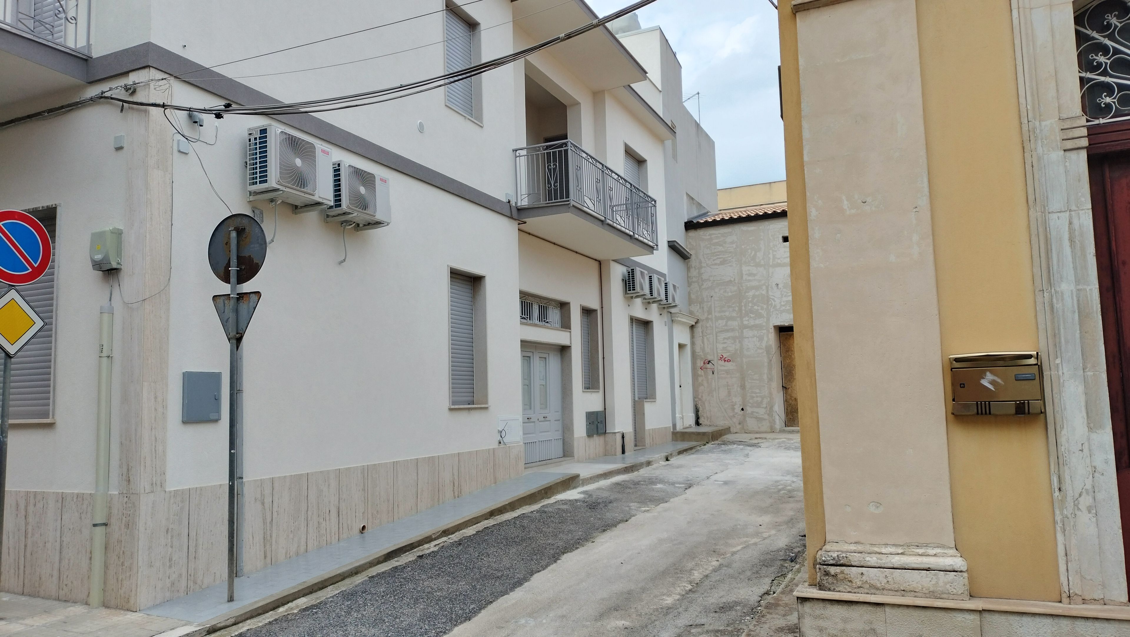 Una calle estrecha flanqueada por edificios, con balcones, una señal de no estacionamiento y un poste de electricidad.