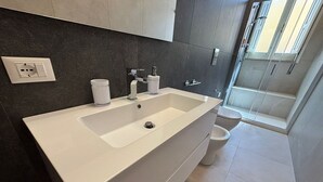 A modern bathroom with a white sink, dark tiled walls, a toilet, and a shower area. - Civico 53 Apartments (Avola)
