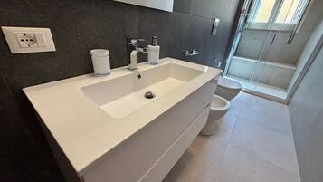 Un bagno moderno con lavandino bianco, pareti piastrellate scure, un water e una doccia.