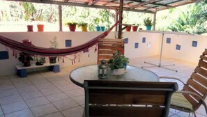 Une terrasse couverte avec une table ronde, des chaises, un hamac, des plantes en pot et un sol carrelé.