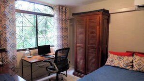 Een slaapkamer met een bed, een bureau met een computer, een kledingkast en een raam met patroon gordijnen.