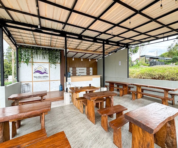 An outdoor dining area with wooden benches and tables, a kitchen counter, and a logo on the wall. - Cozzy Talaga Artea Pangalengan (bandung)