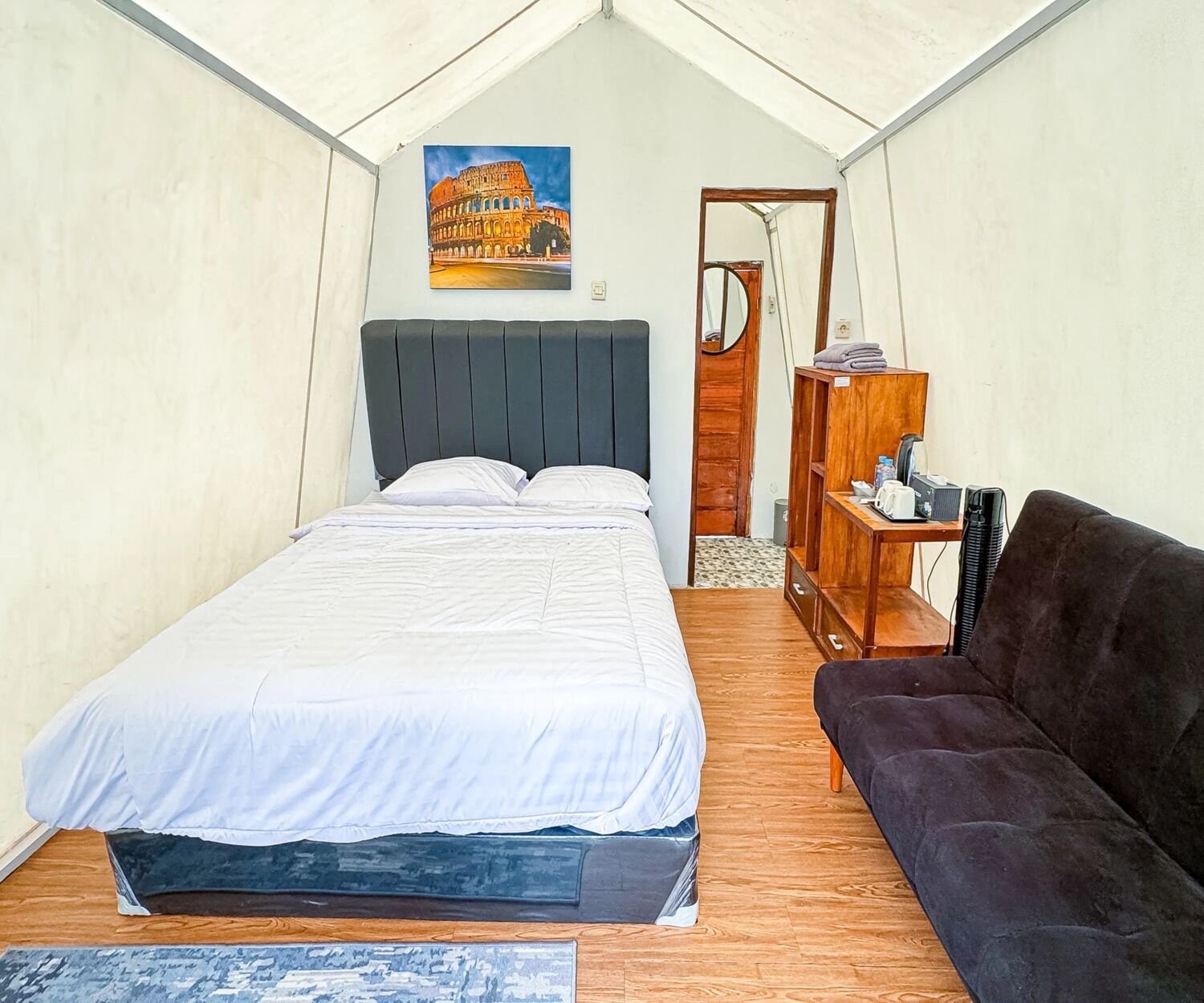 A tented room with a bed, a dark sofa, a wooden side table, and a painting of the Colosseum.