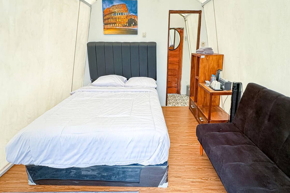 A tented room with a bed, a dark sofa, a wooden side table, and a painting of the Colosseum.