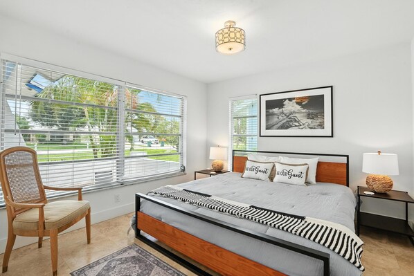 A bedroom with a bed, a chair, a bedside table, a lamp, a framed picture, and large windows with blinds. - New! Beach House - Heated Pool/Spa! 1 Mile to the Beach - Monthly Discounts! (Naples)