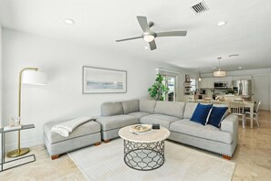 Ein modernes Wohnzimmer mit einer Eckcouch, einem runden Couchtisch, einem Deckenventilator und einer Küche im Hintergrund.