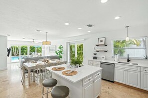 A modern kitchen with white cabinetry, a central island, and a dining area with a pool view. - New! Beach House - Heated Pool/Spa! 1 Mile to the Beach - Monthly Discounts! (Naples)