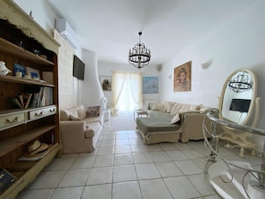 A living room with a sofa, a chair, a bookshelf, a chandelier, and a painting on the wall. - Mykonos Cycladic house
The perfect White House , near panormos beach (Mykonos)