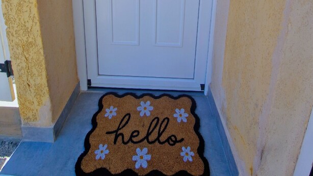Une porte blanche avec une petite fenêtre, un paillasson sur lequel est écrit « bonjour » et orné de fleurs, et un mur jaune.