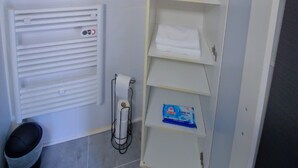 A bathroom with a white radiator, a toilet, a shelf with folded towels, a small plant, and a box of toilet paper.