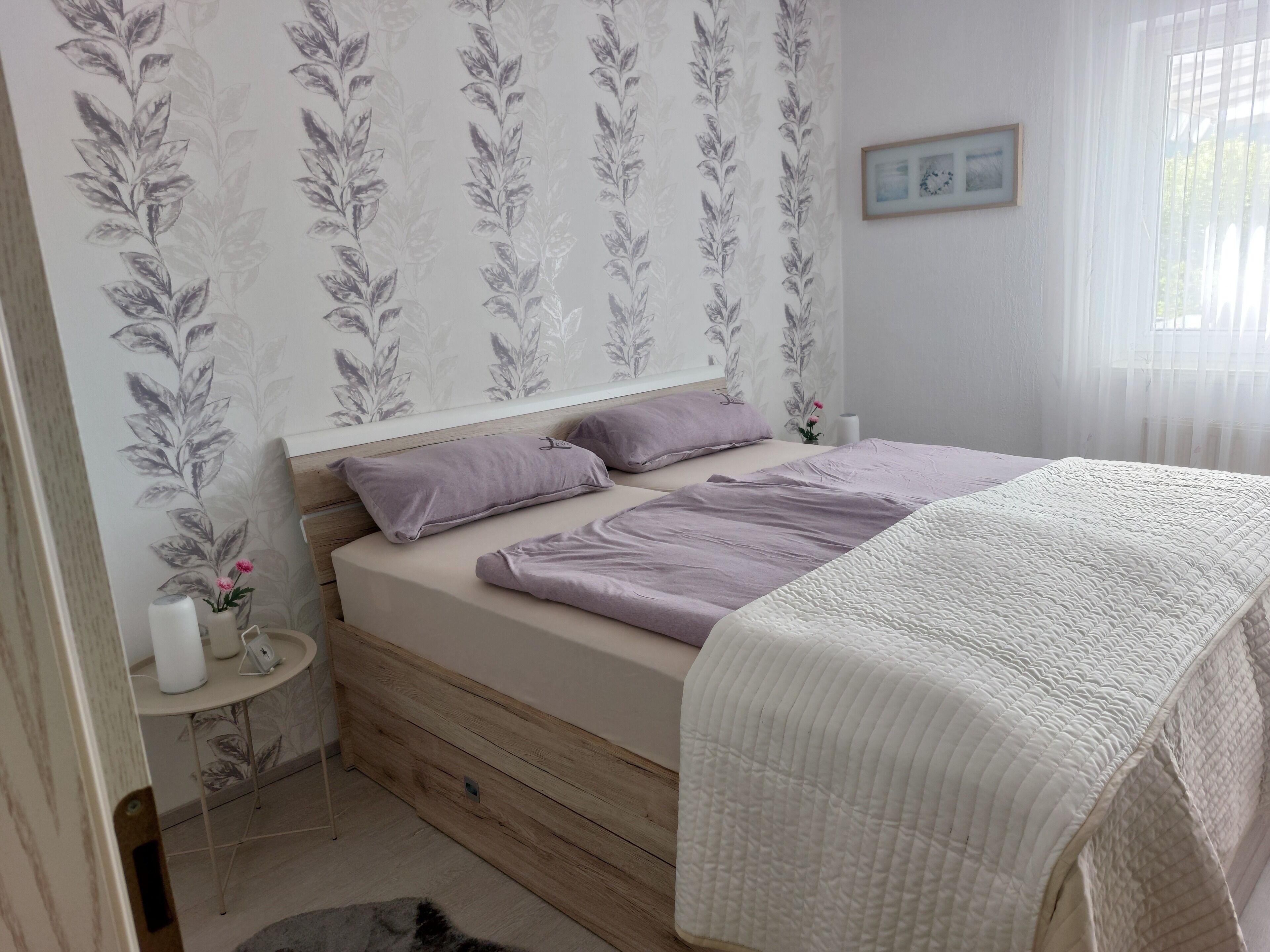 Une chambre à coucher avec un lit, des oreillers, une table de chevet ornée d’un vase, et un mur décoré d’un motif floral.