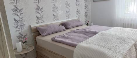 Une chambre à coucher avec un lit, des oreillers, une table de chevet ornée d’un vase, et un mur décoré d’un motif floral.