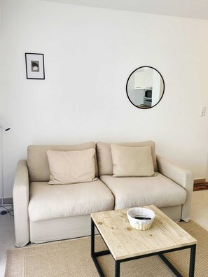 A beige sofa with two cushions, a round mirror on the wall, and a small wooden table with a bowl and a pen. - Comfortable T2 apartment with swimming pool and beach within walking distance (Linguizzetta)