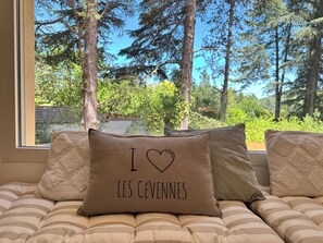 Ein Bett mit gestreifter Bettwäsche und Kissen, eines davon mit dem Aufdruck 'I ♥ LES CEVENNES', und ein Blick aus dem Fenster auf einen Wald.