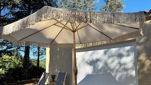 A covered outdoor seating area with a large umbrella, two lounge chairs, and a garage door. - Just a 2-minute walk from downtown Anduze! Enjoy this new home surrounded by nature. (Anduze)