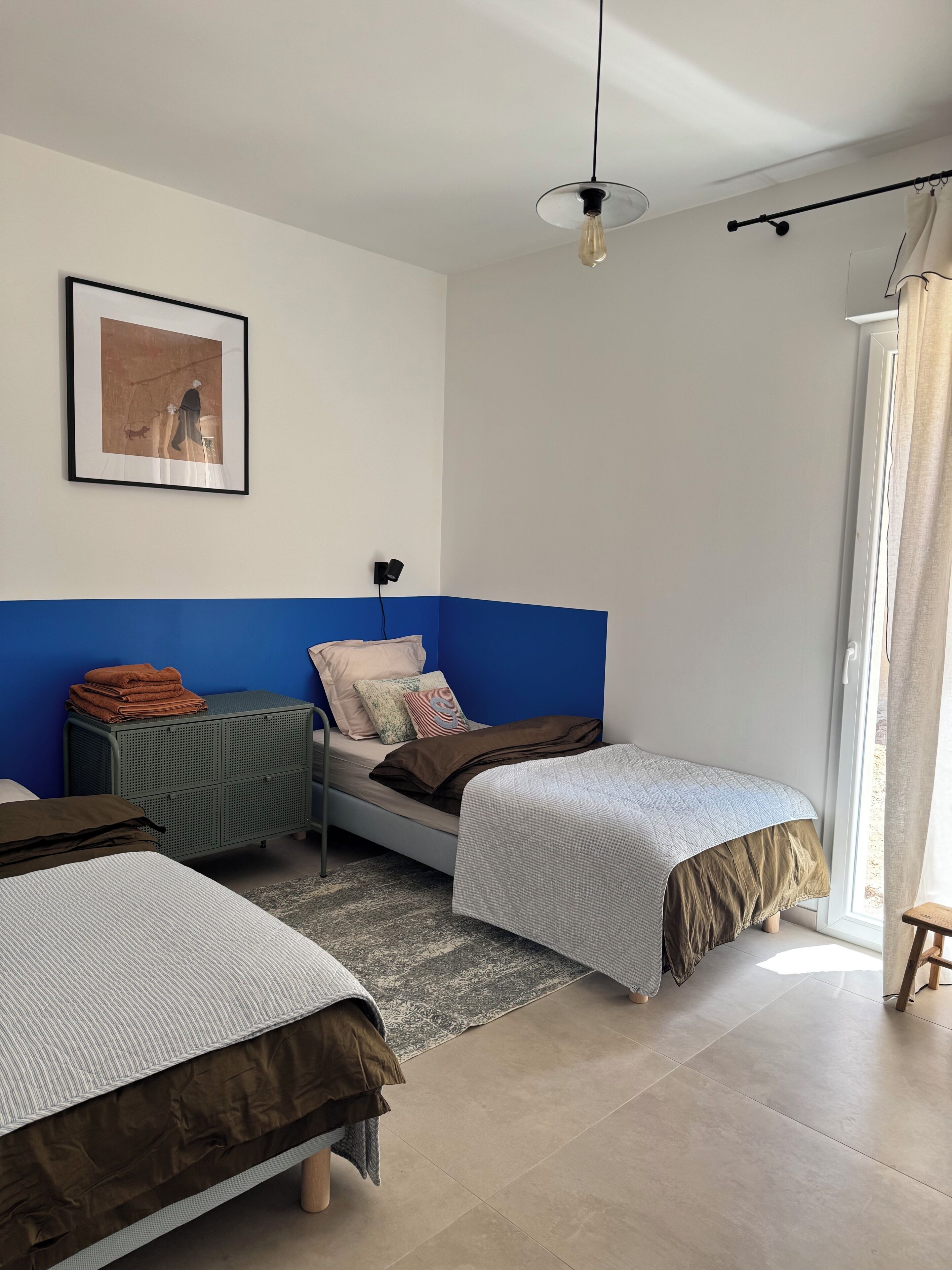 Une chambre avec deux lits, un mur d’accent bleu, un tableau encadré, un petit meuble et une fenêtre donnant sur un paysage.