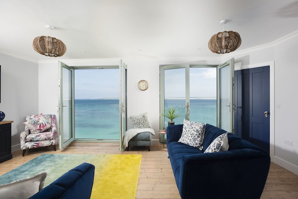 A living room with a sofa, armchair, and a view of the sea through open doors. - Mermaids 2 - Sea views, direct beach access (St Ives)