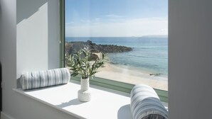 Habitació amb una finestra gran que dona a una platja rocosa i un mar tranquil.