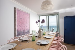 A dining area with a long table, pink chairs, and a view of the sea through large windows. - Mermaids 3 - Sea-facing property, direct beach access (St Ives)