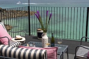A balcony with a view of the sea, a white vase with purple flowers, a wine bottle, and a glass of wine on a glass table. - Mermaids 3 - Sea-facing property, direct beach access (St Ives)