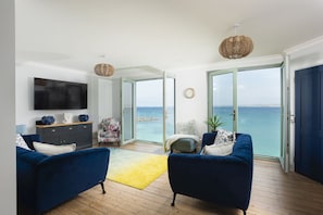 A living room with a TV, two blue sofas, a wooden floor, and a view of the sea through glass doors. - Mermaids - Sea-facing properties, direct beach access (St Ives)
