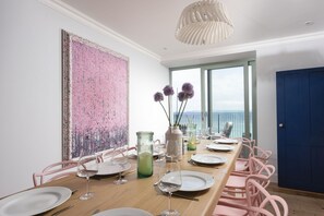 A dining area with a long table, pink chairs, and a view of the sea through large windows. - Mermaids - Sea-facing properties, direct beach access (St Ives)