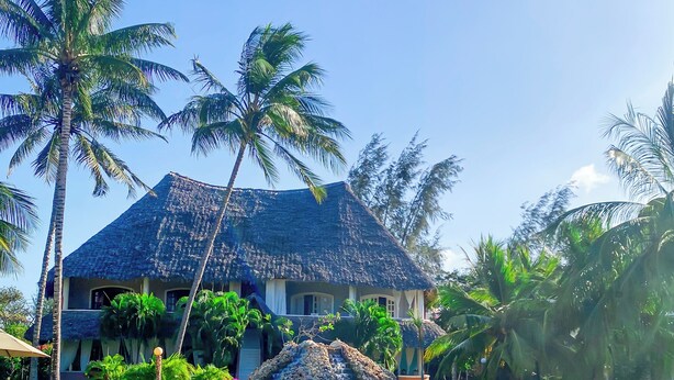 A pool with a wooden lounge chair, a thatched-roof building, and palm trees. - Hidden Gems Homes ocean Beach (Malindi)