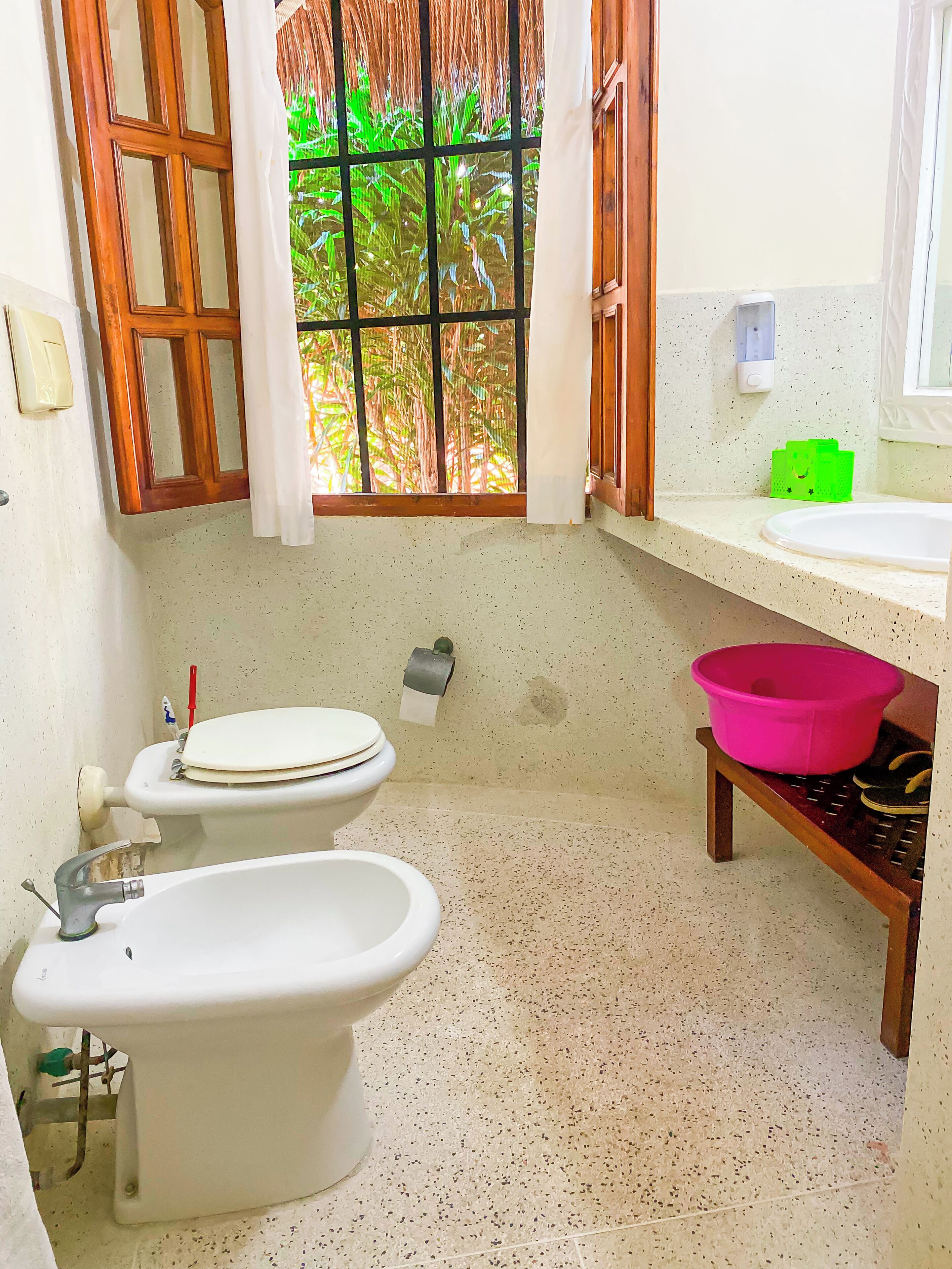 Un baño con dos inodoros, una bañera, un ventanal con vistas a zonas verdes y un estante pequeño con un lavabo rosa.
