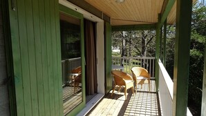 Una terraza de madera con barandilla, un porche cubierto y vista a una casa con ventana y puerta.