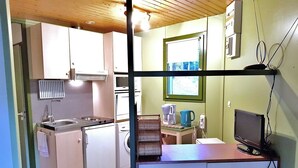 Cocina compacta con lavamanos, estufa y microondas, una pequeña mesa con tetera y cafetera, y un televisor sobre un mueble.