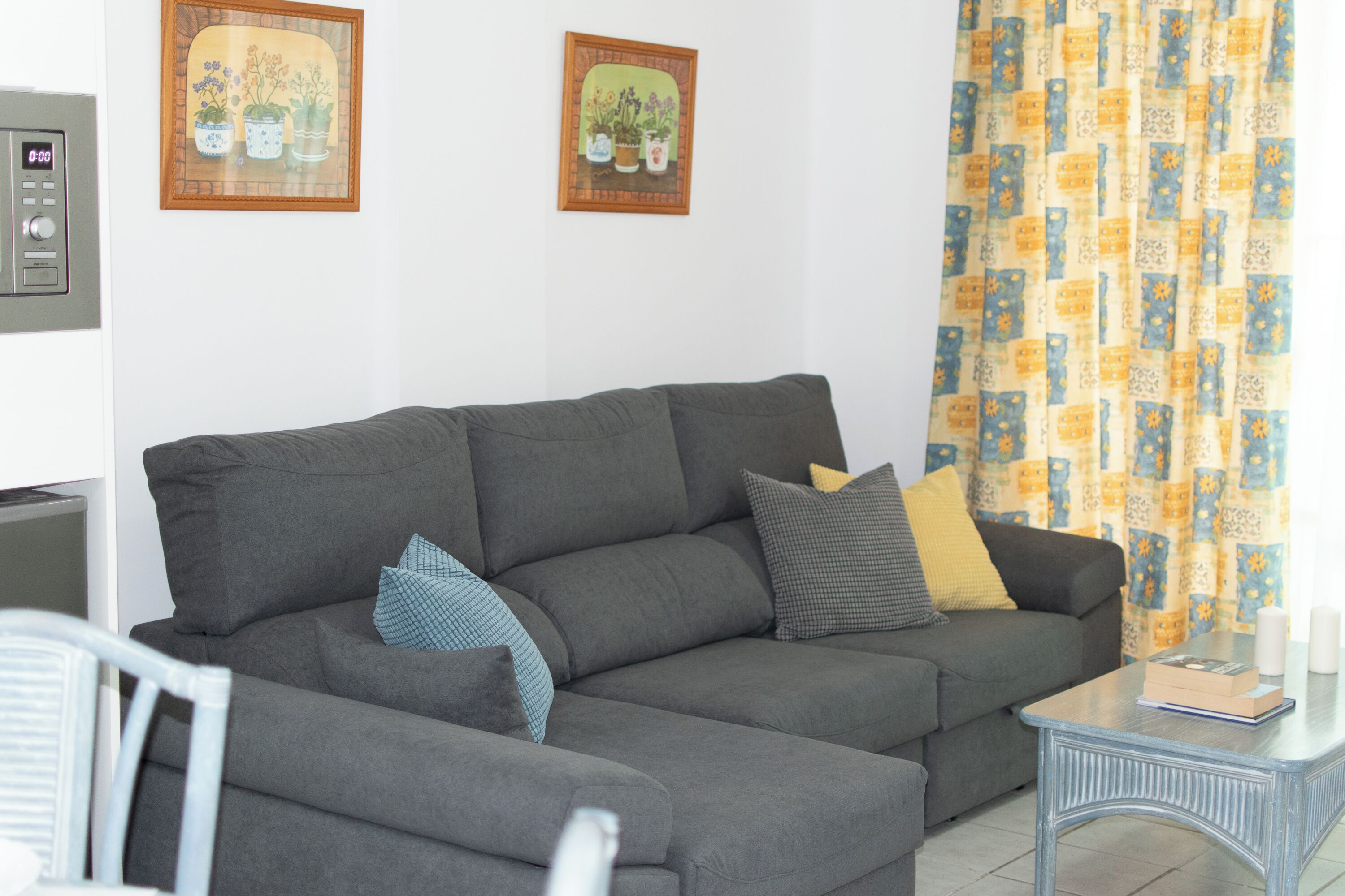 A grey sectional sofa with multiple cushions, a small table with a book, and two framed pictures on the wall.