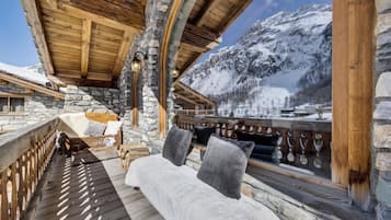 A wooden deck with a stone wall, a bench with cushions, and a view of snow-covered mountains.