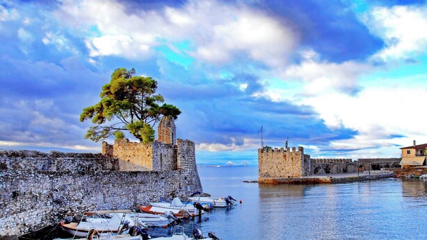 A historic stone wall with a tree, boats docked along the water, and a building with a flag. - In the heart of Nafpaktos's city center, 50m from the sea and 5' from the port. (Nafpaktos)