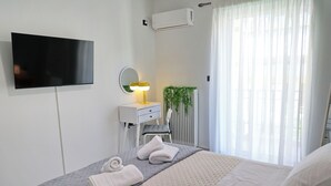 A hotel room with a bed, a TV, a bedside lamp, a mirror, a radiator, and a window with curtains. - In the heart of Nafpaktos's city center, 50m from the sea and 5' from the port. (Nafpaktos)