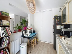 Eine kompakte Küche mit Mikrowelle, Wasserkocher und Kaffeemaschine, ein kleiner Esstisch mit Stühlen, ein Bücherregal und ein Blumentopf.