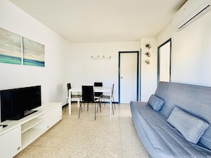 Ein modernes Wohnzimmer mit Flachbildfernseher, grauem Sofa, Esstisch mit Stühlen und einer weißen Wand mit einem Gemälde.