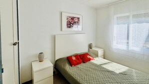 Une chambre à coucher avec un lit, une table de chevet, une commode, un tableau encadré et une fenêtre avec des rideaux.