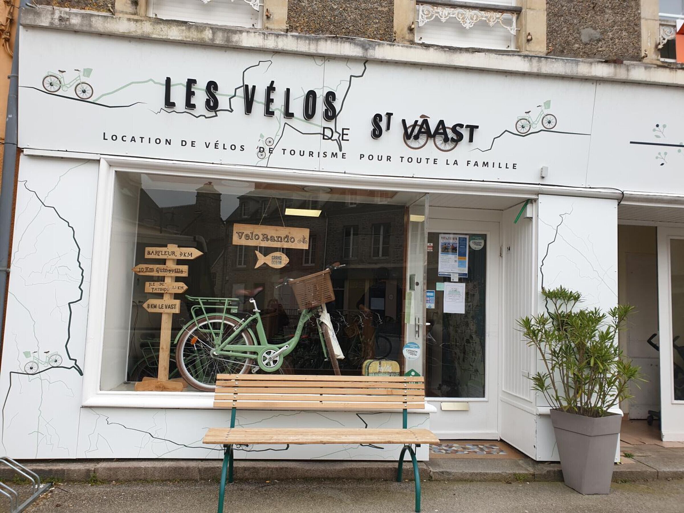 Une devanture de magasin avec une enseigne sur le thème du vélo, un banc à l’extérieur et une plante en pot.