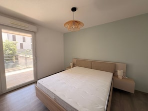 Ein Schlafzimmer mit Bett, Nachttisch, Lampe, Deckenleuchte und einem Fenster mit Blick auf Gebäude und Grünflächen.