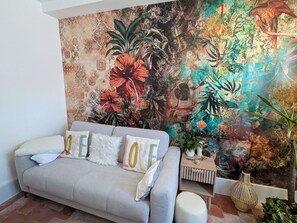 A living room with a sofa, decorative pillows, a small side table, and a large tapestry with a tropical design. - Country house, pets welcome - 6 guests (Tavers)