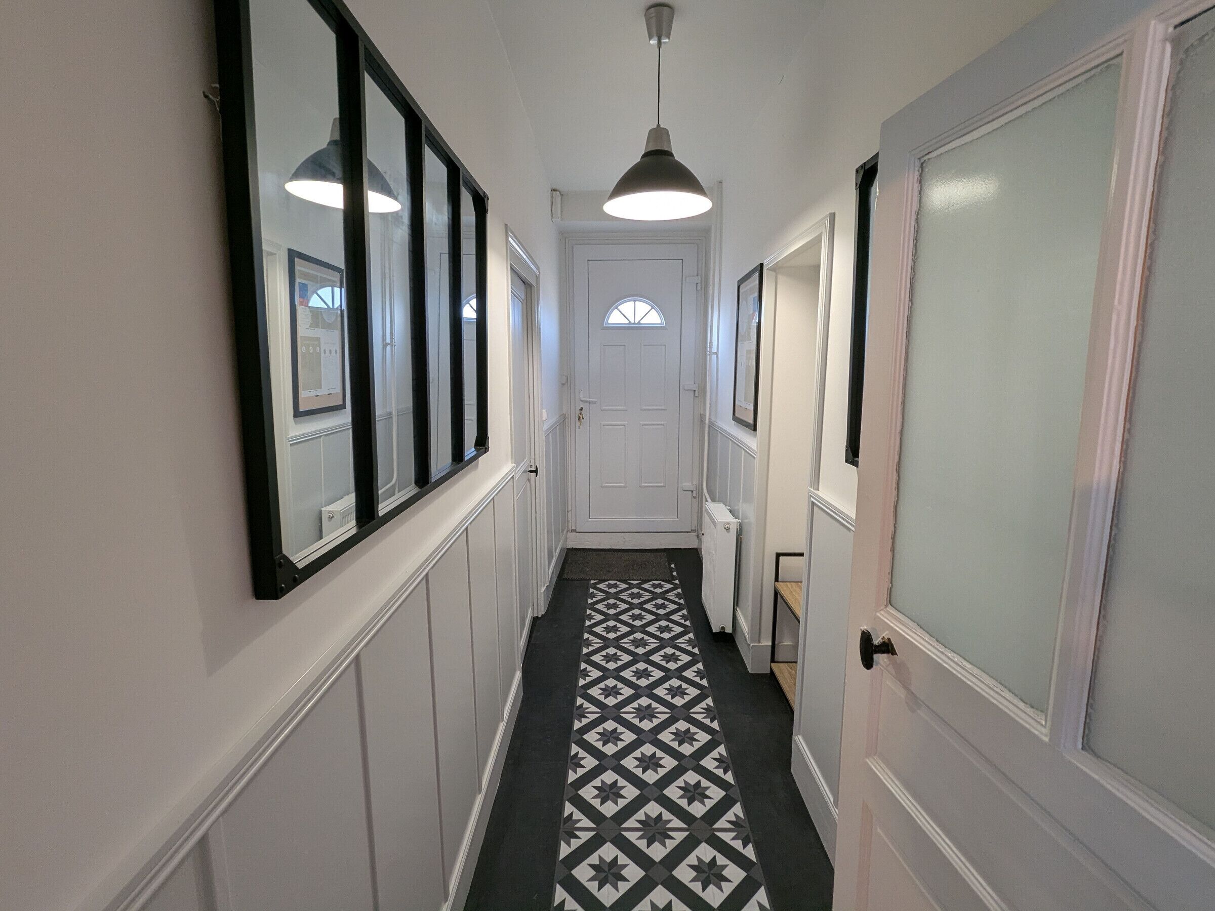 A narrow hallway with a patterned floor, a white door at the end, and a pendant light overhead.
