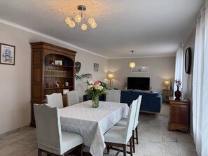 Ein Essbereich mit Tisch, Stühlen, einem Bücherregal, einer Couch, einem Fernseher und einer Zierpflanze.
