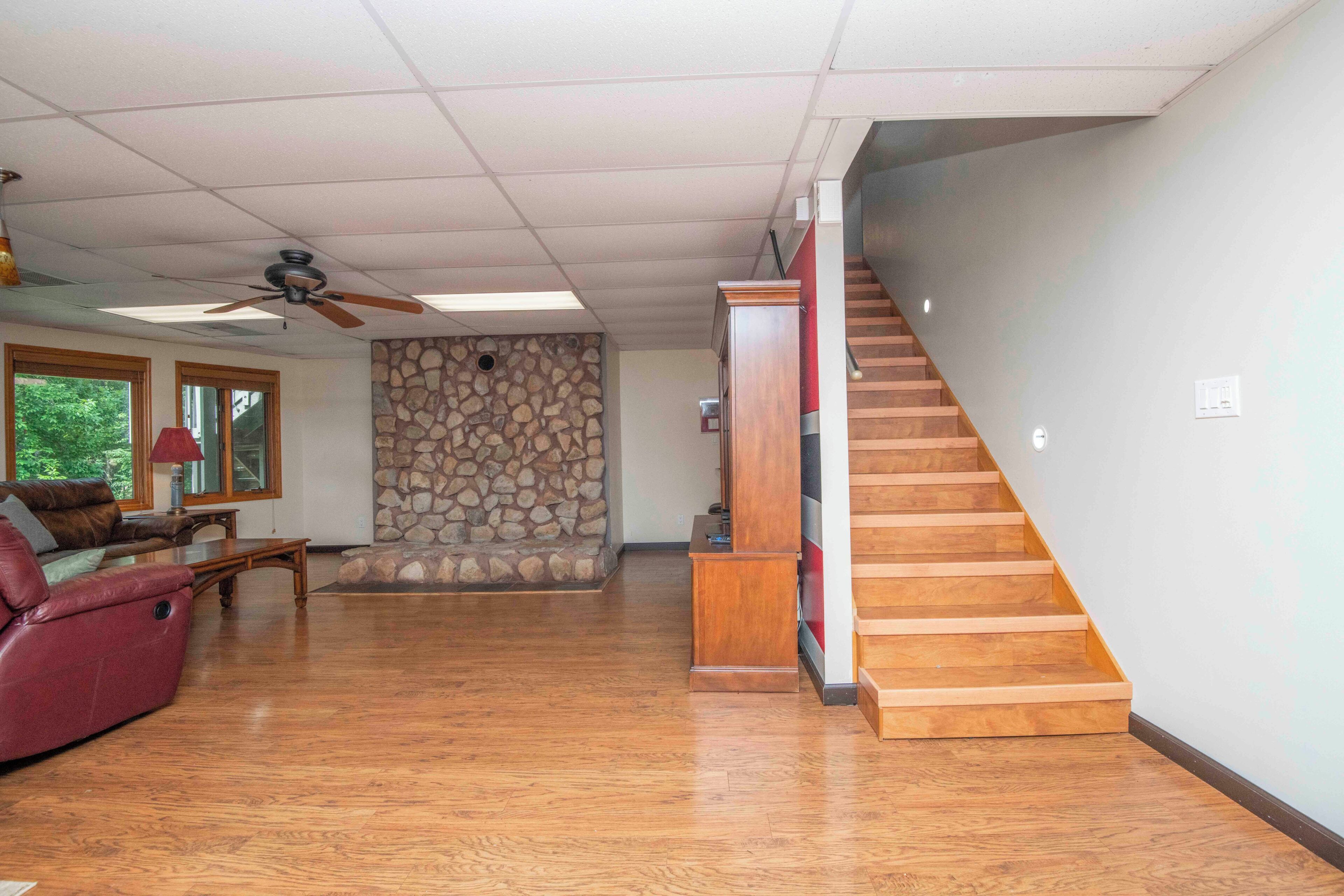 Amplia sala de estar con piso de madera, pared decorativa de piedra, sofá rojo, ventilador de techo y una escalera.