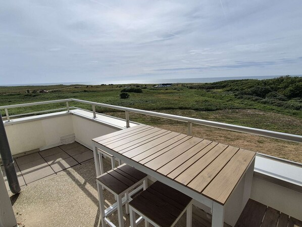 Ein Balkon mit Holztisch und -stühlen, der auf eine grüne Landschaft mit weit entfernten Hügeln und wolkenlosem Himmel blickt.