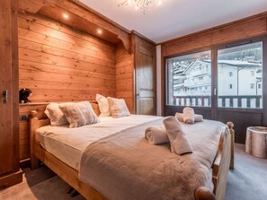 Ein Zimmer mit Holzboden, einem Bett, einem Fenster mit Blick auf Gebäude und einem kleinen Regal mit einem Teddybären.