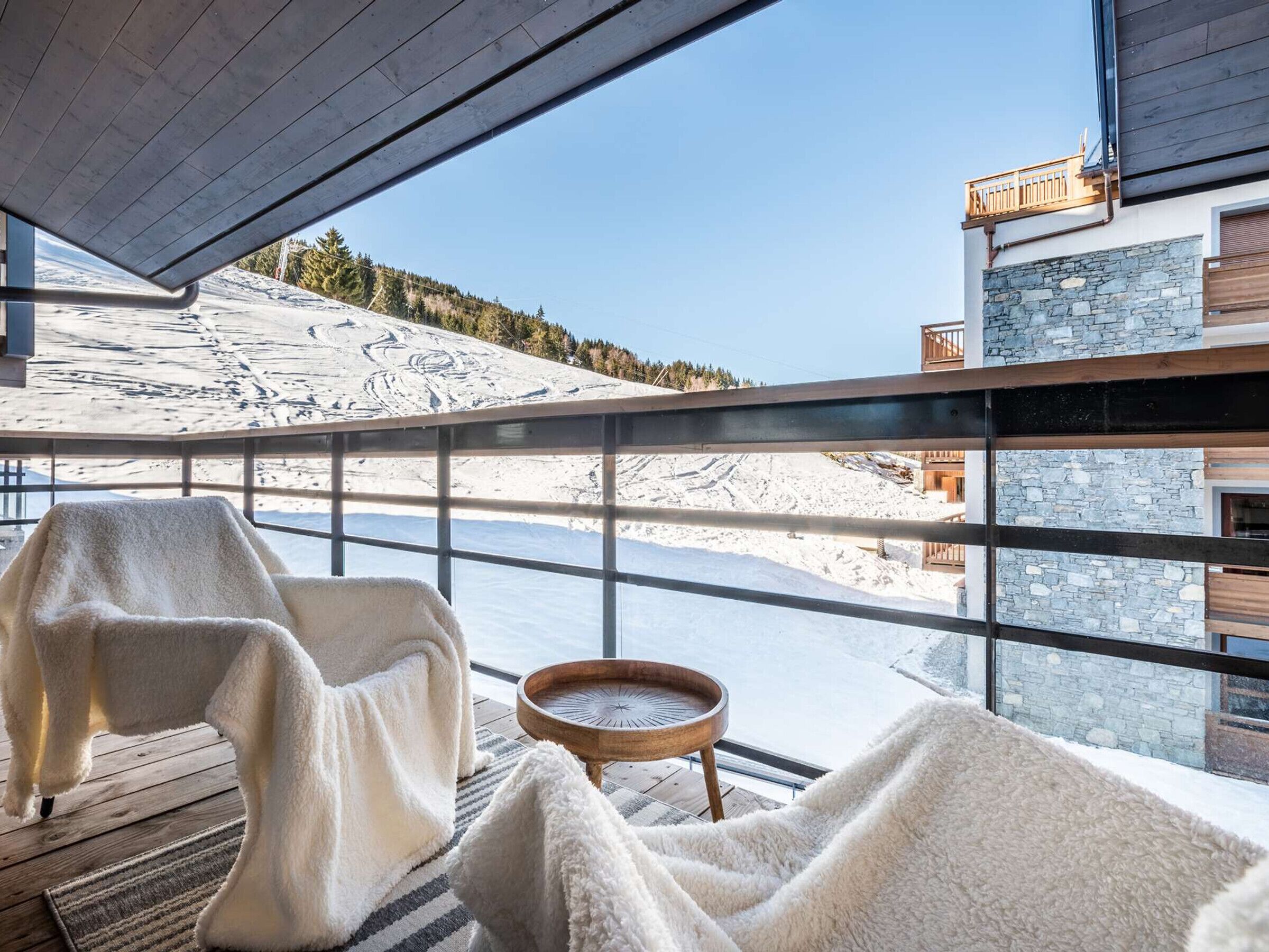 A room with a large window overlooking a snowy landscape, a wooden table, and a chair with a blanket.