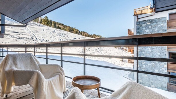 A room with a large window overlooking a snowy landscape, a wooden table, and a chair with a blanket. - Cozy apartment, center station - 4 guests (Courchevel 1550)