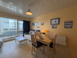 Ein Zimmer mit Esstisch, Stühlen, einer Couch, einem Fenster mit Vorhängen und Bildern an der Wand.