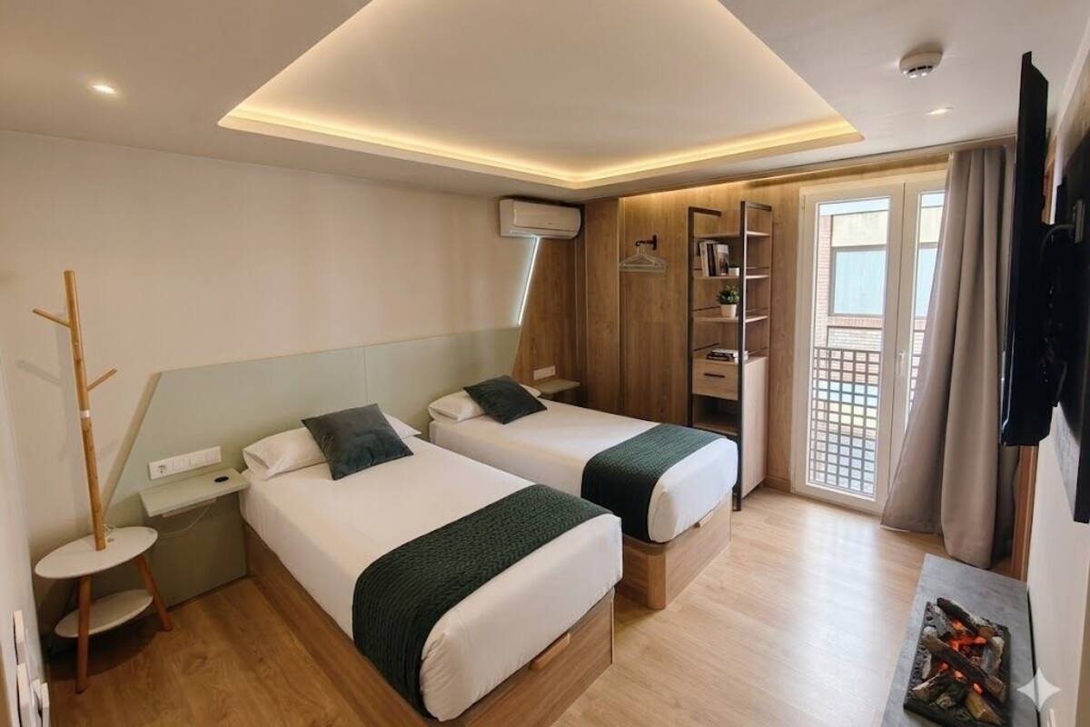 Habitación de hotel con dos camas, una mesita, un armario y un balcón con cortinas.